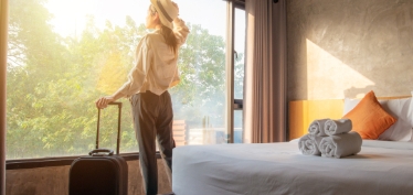 Mujer en habitación de hotel con valija mirando por la ventana.