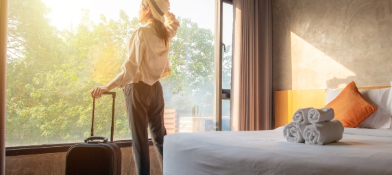Mujer en habitación de hotel con valija mirando por la ventana.